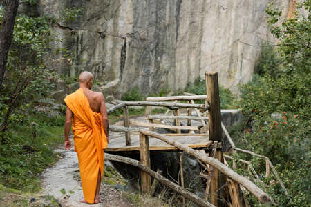 back view of buddhist in orange kasaya walking near wooden walkway and rocks in forestの写真素材