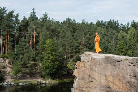 full length view of buddhist in orange robe meditating on rocky cliff over riverの写真素材