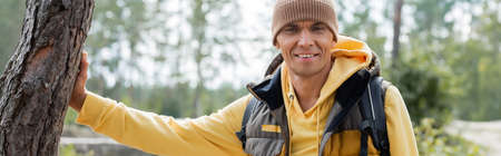 happy tourist in beanie and warm vest looking at camera in forest, bannerの写真素材
