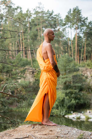 tattooed buddhist in traditional orange robe meditating on hill in forestの写真素材