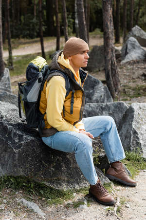 traveler with backpack, wearing warm vest and beanie, sitting on stone in forestの写真素材