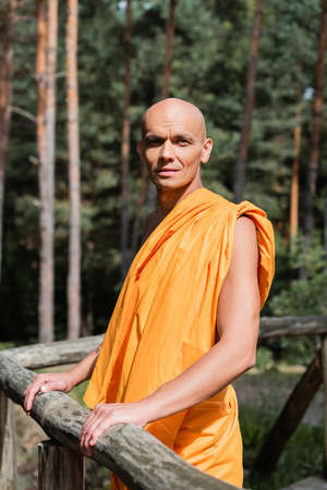 buddhist in orange kasaya looking away while standing near wooden fence in forestの写真素材