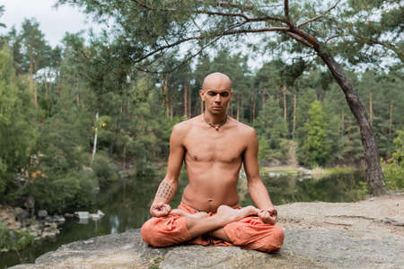 tattooed, shirtless buddhist meditating in lotus pose with closed eyes outdoorsの写真素材
