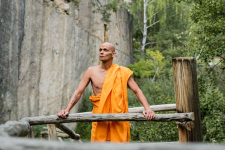 man in traditional buddhist robe looking away near wooden fence and rock in forestの写真素材