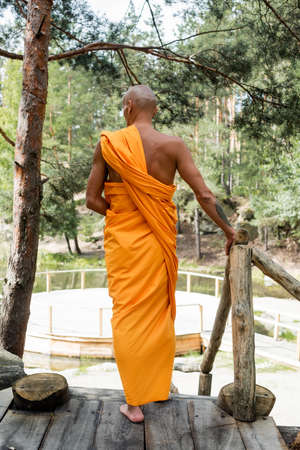 back view of buddhist in traditional orange robe standing near wooden fence in forestの写真素材