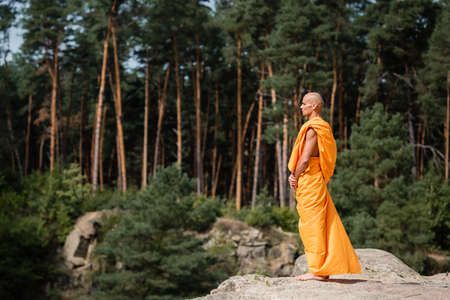 side view of barefoot buddhist monk in orange kasaya meditating on rocky cliff in forestの写真素材