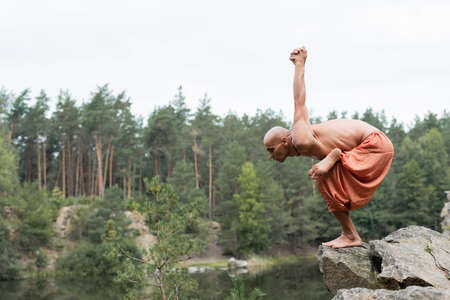 side view of shirtless buddhist meditating in yoga pose on rocky cliff over forest lakeの写真素材