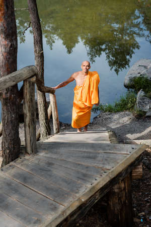 high angle view of buddhist in orange robe walking on wooden stairs near lakeの写真素材