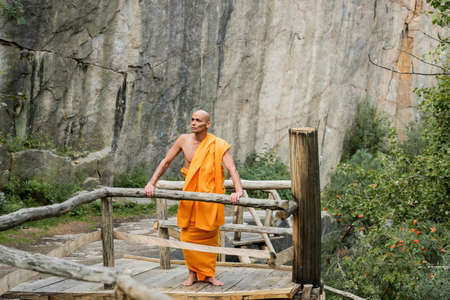 buddhist in orange kasaya standing on wooden walkway near rocks and looking awayの写真素材