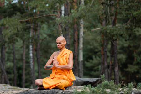buddhist monk in orange kasaya sitting in lotus pose with praying hands while meditating in forestの写真素材