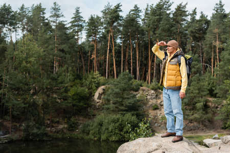full length view of tourist standing on rocky cliff near forest lake and looking awayの写真素材