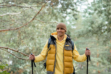 joyful traveler with trekking poles looking away while standing in forestの写真素材