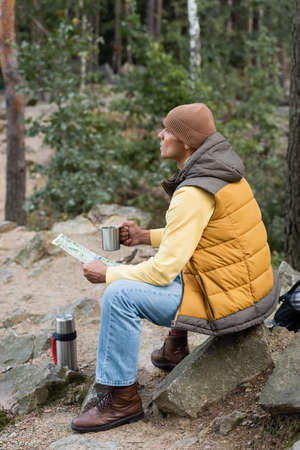 hiker in warm vest and beanie looking away while sitting with mug and map in forestの写真素材