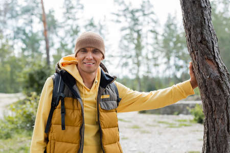 happy traveler in beanie and warm vest smiling at camera in forestの写真素材
