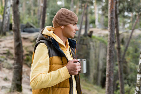 tourist in warm vest and beanie holding metal mug in forestの写真素材