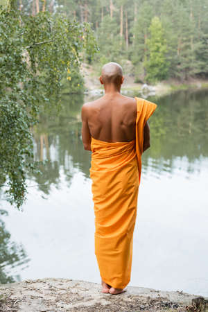 back view of bald man in traditional buddhist robe meditating near forest pondの写真素材