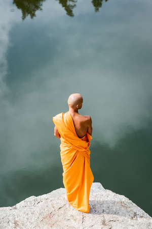 high angle view of buddhist in orange kasaya meditating on rocky cliff over lakeの写真素材
