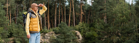 hiker in warm vest and beanie holding hand near forehead and looking away in forest, bannerの写真素材