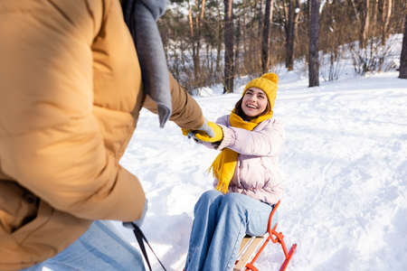 Cheerful woman holding hand of boyfriend while riding sleigh in winter parkの写真素材