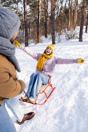 Cheerful woman sitting on sled near blurred boyfriend in winter parkの写真素材