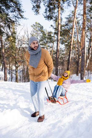 Smiling man pulling sleigh with girlfriend in winter parkの写真素材