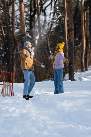 Side view of excited couple in hats throwing snow near sled in parkの写真素材