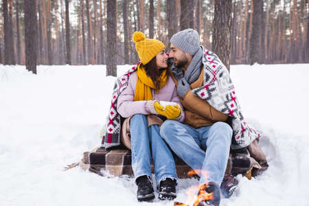 Side view of positive couple with cup and blanket looking at each other near blurred bonfire in winter parkの写真素材