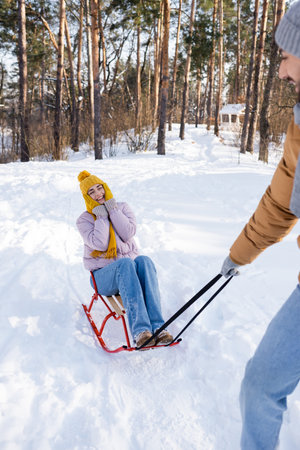 Cheerful woman in gloves and hat sitting on sleigh near blurred boyfriend in winter parkの写真素材