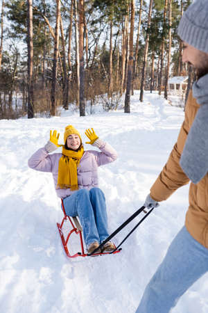 Happy woman waving hands while sitting on sled near blurred boyfriend in winter parkの写真素材