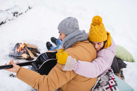 High angle view of woman hugging boyfriend playing acoustic guitar near bonfire in winter parkの写真素材