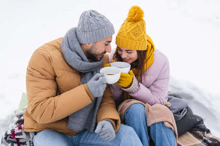 Positive couple holding cups of coffee sitting on blankets on snowの写真素材