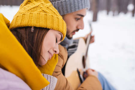 Side view of woman sitting near blurred boyfriend with acoustic guitar in winter parkの写真素材
