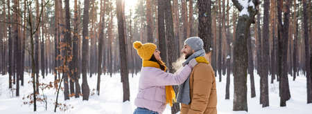 Side view of cheerful woman embracing boyfriend in hat and scarf in winter park, bannerの写真素材