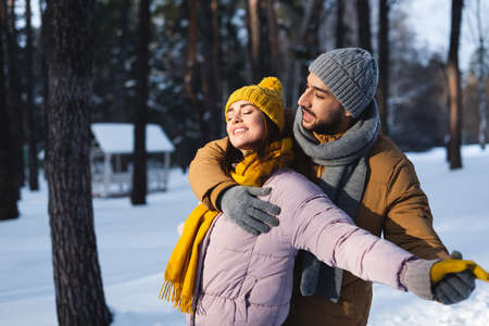 Man hugging and holding hand of happy girlfriend with closed eyes in winter parkの写真素材