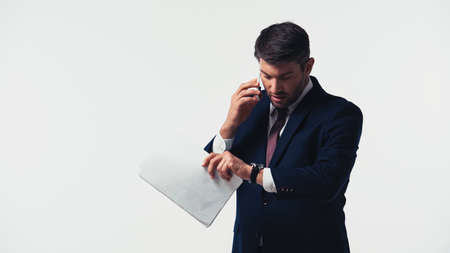 Businessman talking on smartphone while holding papers and looking at wristwatch isolated on whiteの写真素材