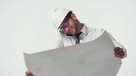 Smiling engineer talking on smartphone and holding blurred blueprint isolated on whiteの写真素材
