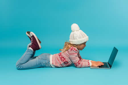side view of girl in hat, sweater and jeans typing on laptop with blank screen on blue