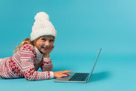 cheerful girl in knitted hat and sweater with ornament using laptop while lying on blueの写真素材