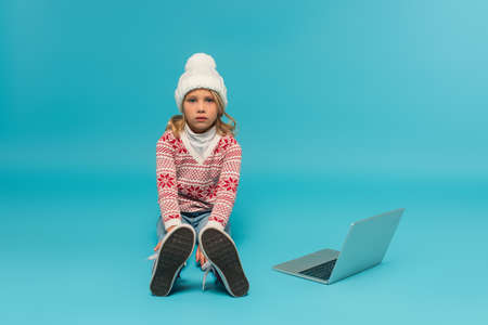child in knitted hat and jumper with ornament sitting near laptop on blueの写真素材