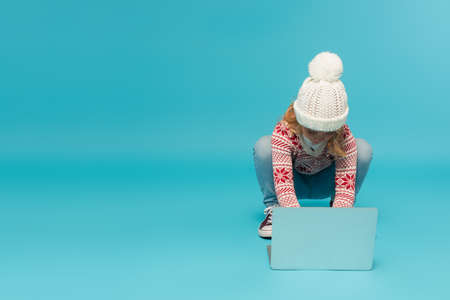 child in warm hat and sweater using laptop on blueの写真素材