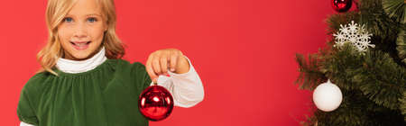 smiling child holding shiny christmas ball near decorated spruce isolated on red, bannerの写真素材