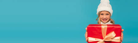 joyful child in warm hat smiling at camera while holding present isolated on blue, bannerの写真素材