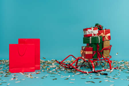 sled with multicolored gift boxes near red shopping bags on blue