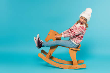 amazed kid in sweater and hat screaming while riding rocking horse on blueの写真素材