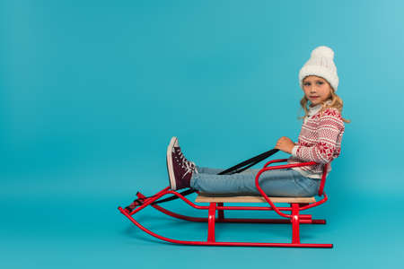 kid in knitted hat and sweater sitting in sled and looking at camera on blueの写真素材
