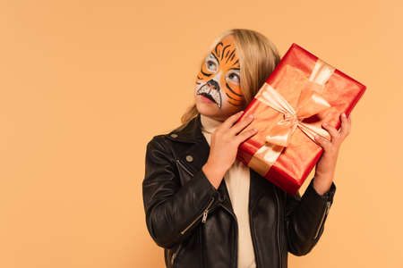 curious child in black leather jacket and tiger makeup holding present isolated on beigeの写真素材