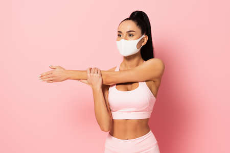 young sportswoman in medical mask warming up on pink