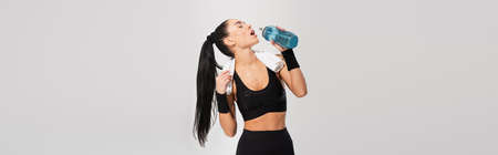 young sportswoman with towel on shoulders drinking water isolated on gray, banner