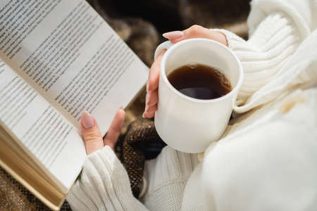 cropped view of woman reading book while holding cup of warm teaの写真素材