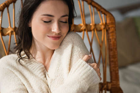 pleased woman in warm sweater hugging herself while resting at home with closed eyesの写真素材